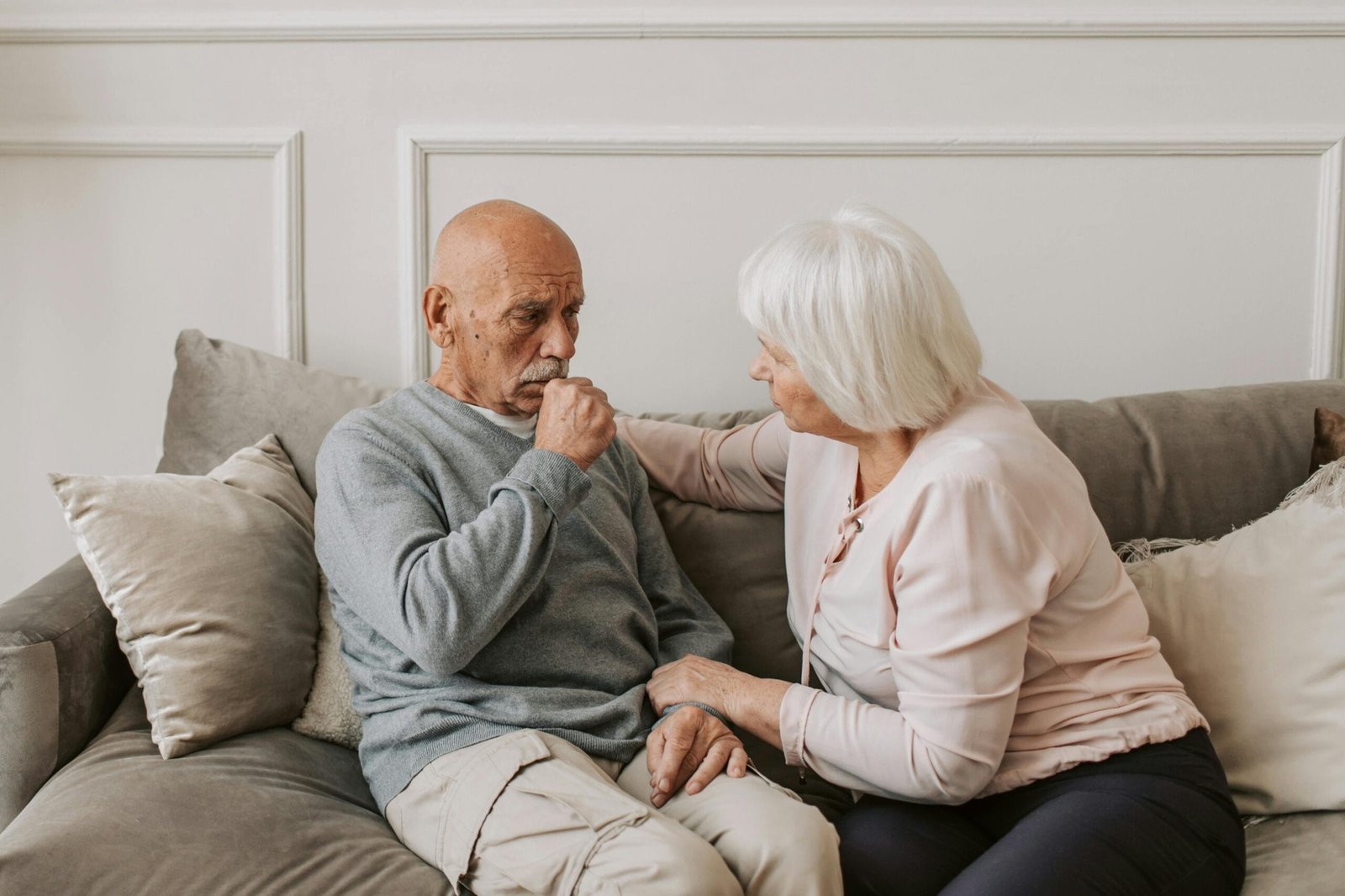 An elderly woman comforts a man coughing on a couch, showcasing care and affection.